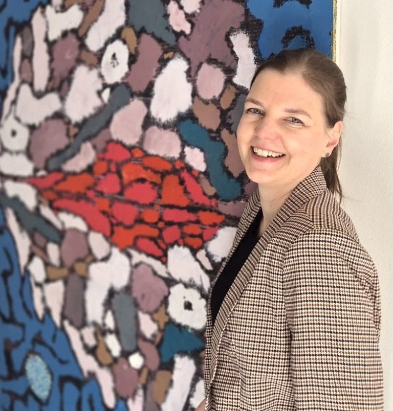 Verena Wessel steht im Jeanshemd vor einer Wand und lächelt.